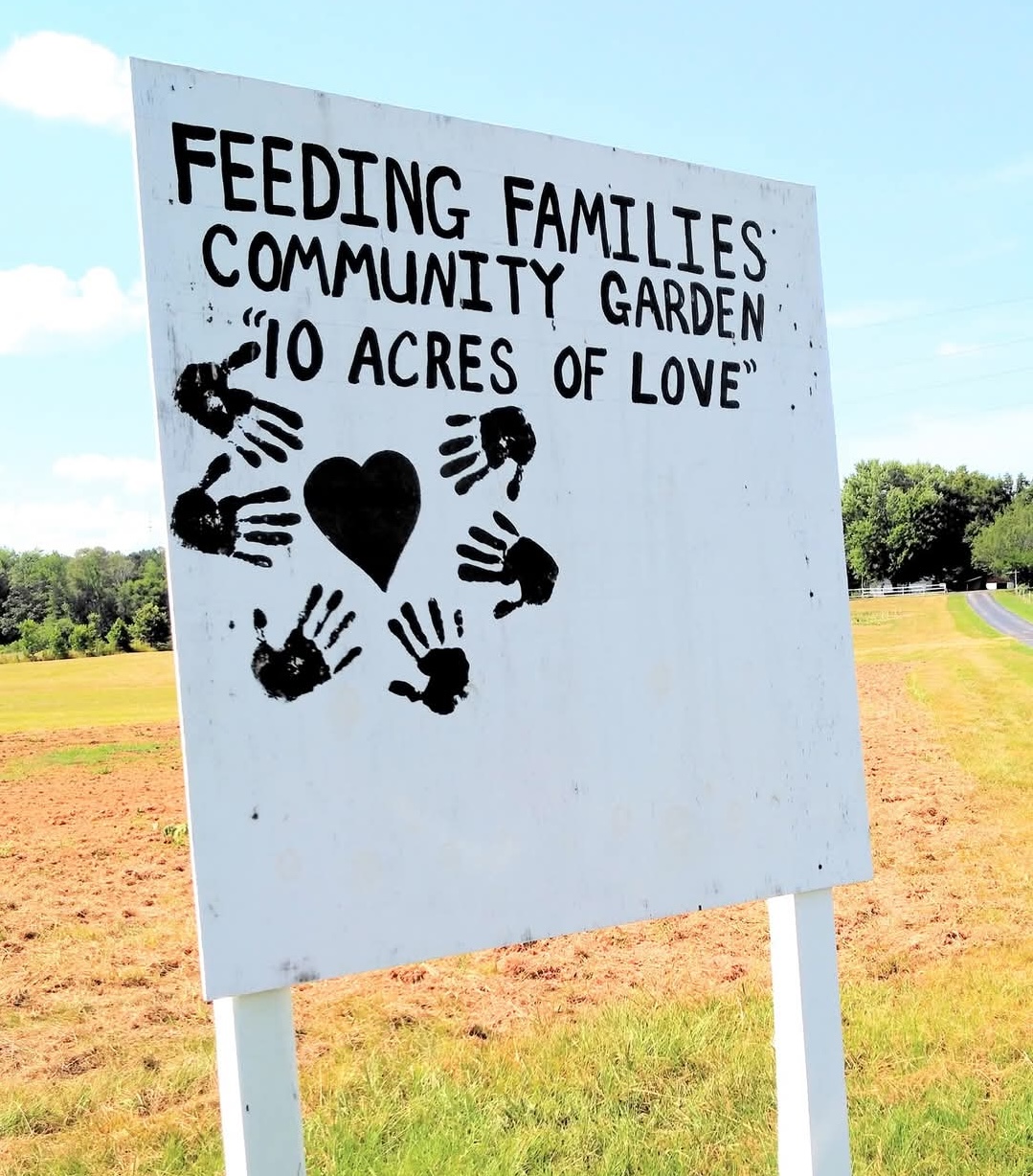 A white wooden sign with the text "Feeding families community garden- 10 acres of love" as well as hand prints and a heart symbol. The background is a open green field with a treeline and a bright blue sky.